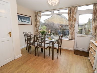 A dining area with a glass table and six chairs near a window and door with floral curtains at Rosemary Cottage in Bourton-on-the-Water