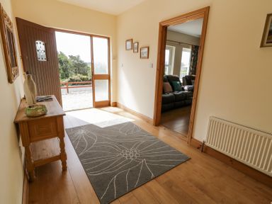An entrance hallway with a wooden side table vase and decorative item on it a gray rug with a floral pattern a door open to outside and a view of a living room with dark sofas at Creeslough View Creeslough near Glenveagh National Park County Donegal