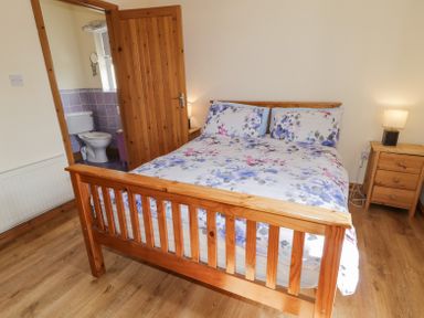 A bedroom with a wooden double bed and floral bedding next to a wooden nightstand with a lamp and an open door showing a bathroom with a toilet at Creeslough View in Creeslough near Glenveagh National Park County Donegal