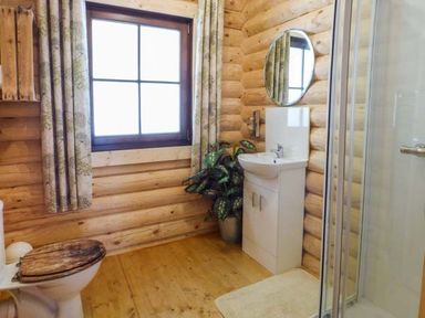 A bathroom with a wooden toilet seat a white sink a round mirror a window with curtains and a shower at Wilderness Lodge in Stiperstones