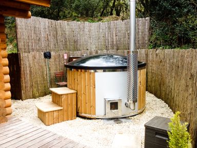 An outdoor wooden hot tub with a metal chimney and steps surrounded by a reed fence at Wilderness Lodge in Stiperstones