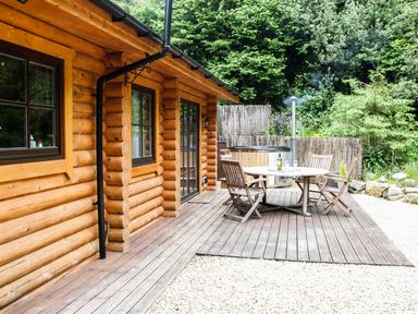 Wooden patio with table and chairs next to log cabin surrounded by trees at Wilderness Lodge in Stiperstones