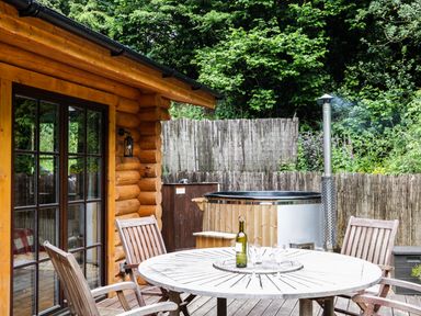 A wooden patio with a round table and four chairs outside a log cabin with a hot tub and greenery in the background at Wilderness Lodge in Stiperstones