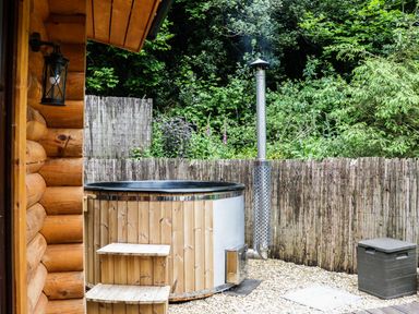 An outdoor hot tub next to a wooden cabin with a metal chimney and a plastic storage box at Wilderness Lodge in Stiperstones