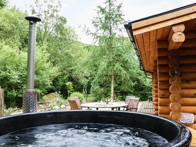 An outdoor patio with a black hot tub round wooden table and chairs beside a log cabin at Wilderness Lodge in Stiperstones