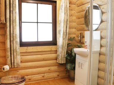 A bathroom with wooden log walls a window with curtains a white sink a round mirror a plant and a wooden toilet seat at Wilderness Lodge in Stiperstones