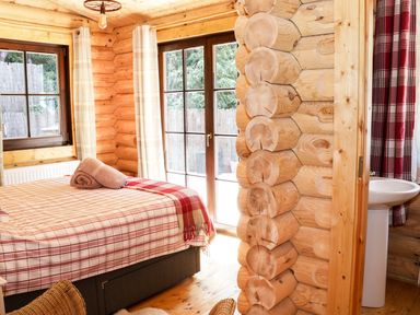 A bedroom with a bed covered in plaid bedding and towels near windows and log walls at Wilderness Lodge in Stiperstones
