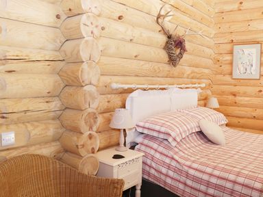 A bedroom with a checkered bedspread a wicker chair bedside lamps and wooden log walls at Wilderness Lodge in Stiperstones