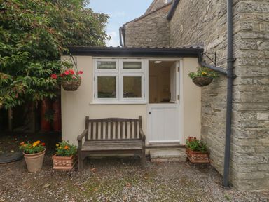 A small outdoor area with a wooden bench flowers in pots hanging baskets and a door and window on a stone wall at Drovers in Sutton Mallet
