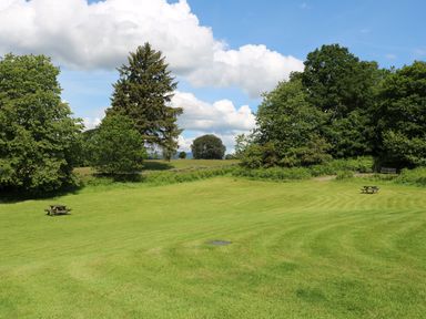 A large grassy field with two wooden picnic tables and trees in the background at Buttermere Sawrey near Hawkeshead