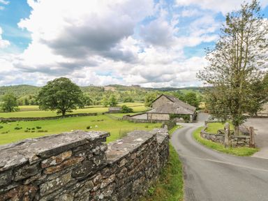 A narrow paved road leading to stone buildings surrounded by grassy fields and trees at Buttermere in Sawrey near Hawkeshead