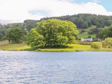 A lake with two people in a boat near an island with trees and a countryside landscape with houses and hills in the background at Buttermere Sawrey near Hawkeshead