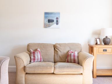 A beige sofa with two red and white plaid pillows a wooden side table with a lamp and photo frame and a small wall art above the sofa at Buttermere Sawrey near Hawkeshead