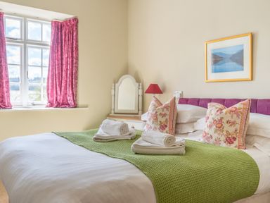 A bedroom with a double bed a green throw folded towels floral pillows a window with pink curtains and a framed picture at Buttermere Sawrey near Hawkeshead