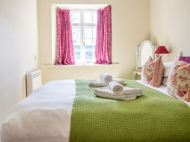 A bedroom with a bed covered by a green blanket and white towels near a window with pink curtains and a side table with a lamp and mirror at Buttermere Sawrey near Hawkeshead