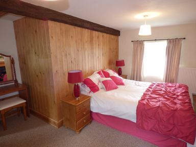 A bedroom with a bed with white and red bedding wooden nightstands with red lamps a wooden vanity with stool and a window with curtains at Ffynnon Gower in Llangower near Bala