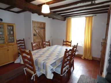 A dining room with a table covered by a patterned tablecloth surrounded by wooden chairs by a window with yellow curtains at Ffynnon Gower in Llangower near Bala