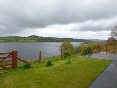 A garden with grass and stone patio overlooking a lake with hills and trees in the background at Ffynnon Gower in Llangower near Bala
