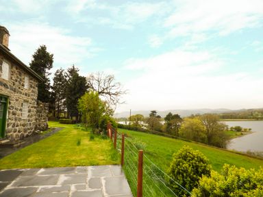 A stone house with a green door beside a lawn and a fence overlooking a lake and trees at Ffynnon Gower in Llangower near Bala