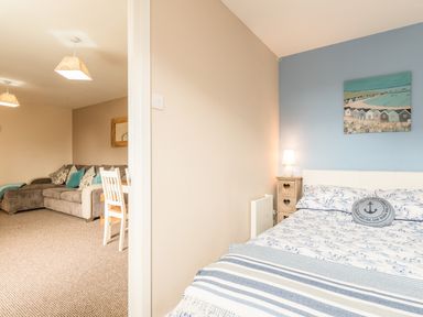 A bedroom with a bed and bedside lamp next to an open doorway showing a dining area with a sofa at Rock Rose in Beadnell