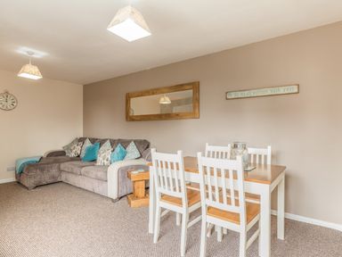 A living room with a dining table and chairs a sofa with cushions a wall mirror and a clock at Rock Rose in Beadnell