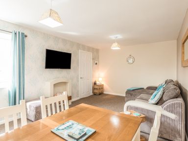 A living room with a dining table, chairs, a wall-mounted TV, sofa, small side table with lamp, and patterned wallpaper at Rock Rose in Beadnell