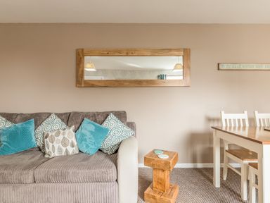 A living room with a beige sofa with blue and patterned cushions a wooden side table a dining table with white chairs and a wooden framed mirror on the wall at Rock Rose in Beadnell