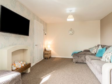 A living room with a grey sofa and blue cushions a small lamp on a side table a wall clock and a flat screen tv above a fireplace at Rock Rose in Beadnell