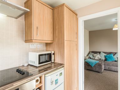 A kitchen with wooden cabinets a microwave a stovetop and a view of a living room with a sofa and cushions at Rock Rose in Beadnell