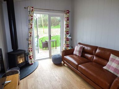 A living room with a brown leather sofa two pillows a wood burning stove a sliding glass door with curtains and a small outdoor table and chair at Shepherd Lodge in Allithwaite near Grange-over-Sands