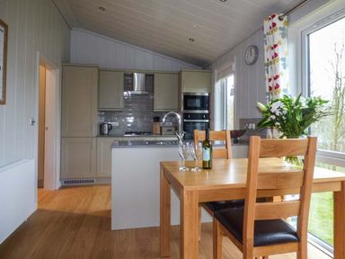 A kitchen and dining area with wooden table and chairs flowers on the table and kitchen cabinets at Shepherd Lodge in Allithwaite near Grange-over-Sands