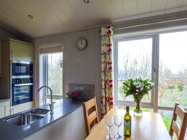 A kitchen with a sink, oven, dining table, wine bottle, wine glasses, flowers, and windows with floral curtains at Shepherd Lodge in Allithwaite near Grange-over-Sands