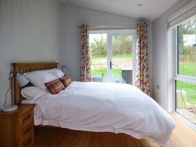 A bedroom with a wooden bed and white bedding next to glass doors with colorful curtains at Shepherd Lodge in Allithwaite near Grange-over-Sands