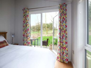 A bedroom with a bed a nightstand and a lamp next to glass doors with floral curtains at Shepherd Lodge in Allithwaite near Grange-over-Sands