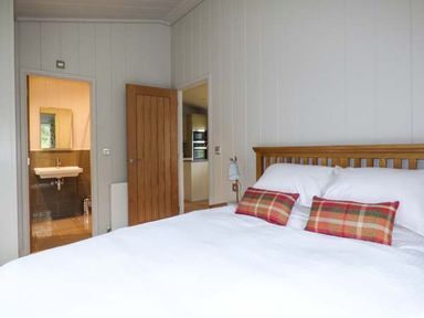 A bedroom with a wooden bed frame white bedding and plaid pillows with a door leading to a bathroom and another door leading to a kitchen at Shepherd Lodge in Allithwaite near Grange-over-Sands
