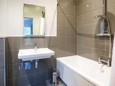 A bathroom with a wall-mounted sink a mirror a bathtub with a showerhead and tiled walls at Shepherd Lodge in Allithwaite near Grange-over-Sands