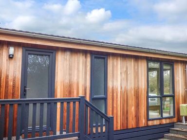 The exterior of a wooden cabin with dark framed doors and windows and a small porch with railings at Shepherd Lodge in Allithwaite near Grange-over-Sands