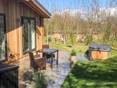 A patio with a table and two chairs next to a wooden building with large windows and a hot tub in the garden at Shepherd Lodge in Allithwaite near Grange-over-Sands