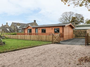 A wooden single-story house with a fenced yard and gravel driveway at Thorntree Lodge in Rowsley