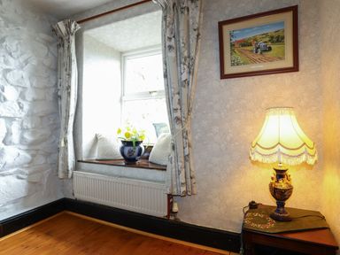 A window with curtains and cushions next to a table with a lamp beneath a framed picture at Hendy Cottage in Llanbedr near Harlech