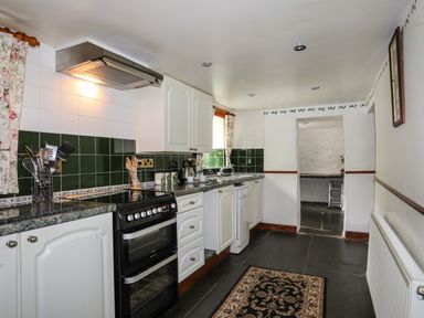 A kitchen with white cabinets green tiles a black oven utensils on the counter a rug on the floor and a window at Hendy Cottage in Llanbedr near Harlech