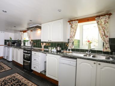 A kitchen with white cabinets green tiled backsplash a black stove and a window with floral curtains at Hendy Cottage in Llanbedr near Harlech