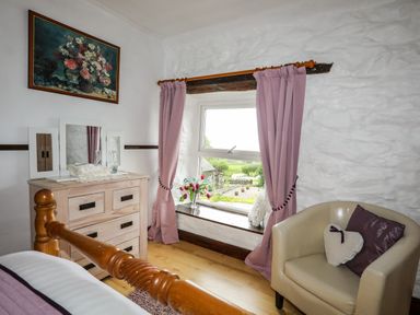 A bedroom corner with a wooden dresser and mirror pink curtains on a window and a beige chair with cushions at Hendy Cottage in Llanbedr near Harlech
