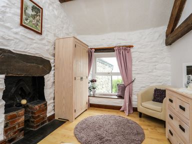 A room with a window and pink curtains a wooden wardrobe a beige armchair and a fireplace at Hendy Cottage in Llanbedr near Harlech