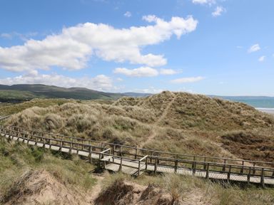 A wooden walkway through sand dunes with grass near a beach and hills in the background at Hendy Cottage Llanbedr near Harlech