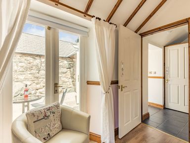 A room with a white armchair and floral cushion near glass doors leading to an outdoor area with a stone wall and a small table with a bottle at Y Becws in Llanbedr near Harlech