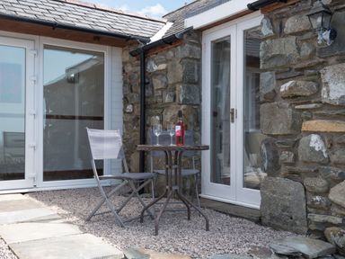 A small outdoor seating area with a round table and two chairs against a stone wall with glass doors at Y Becws in Llanbedr near Harlech