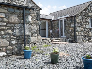 An outdoor area with stone buildings a small table and chair set and potted plants on gravel at Y Becws in Llanbedr near Harlech