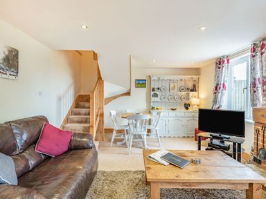 A living room with a brown leather sofa a wooden coffee table a dining table with white chairs a white cabinet a TV and stairs at Harvest Moon in Grundisburgh