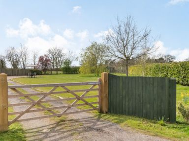 A wooden gate opening to a gravel driveway with a lawn and trees beyond at Harvest Moon in Grundisburgh